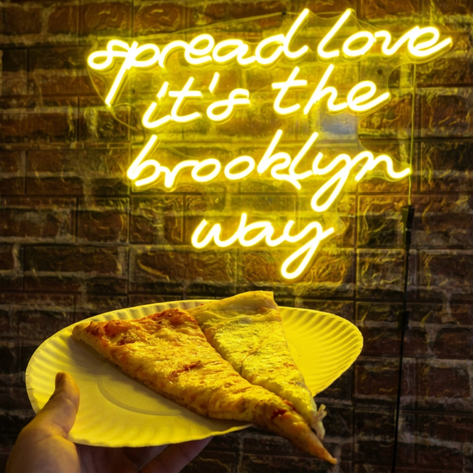 Two slices of pizza on a paper plate held before a brick wall with a glowing “spread love, it’s the Brooklyn way” sign.
