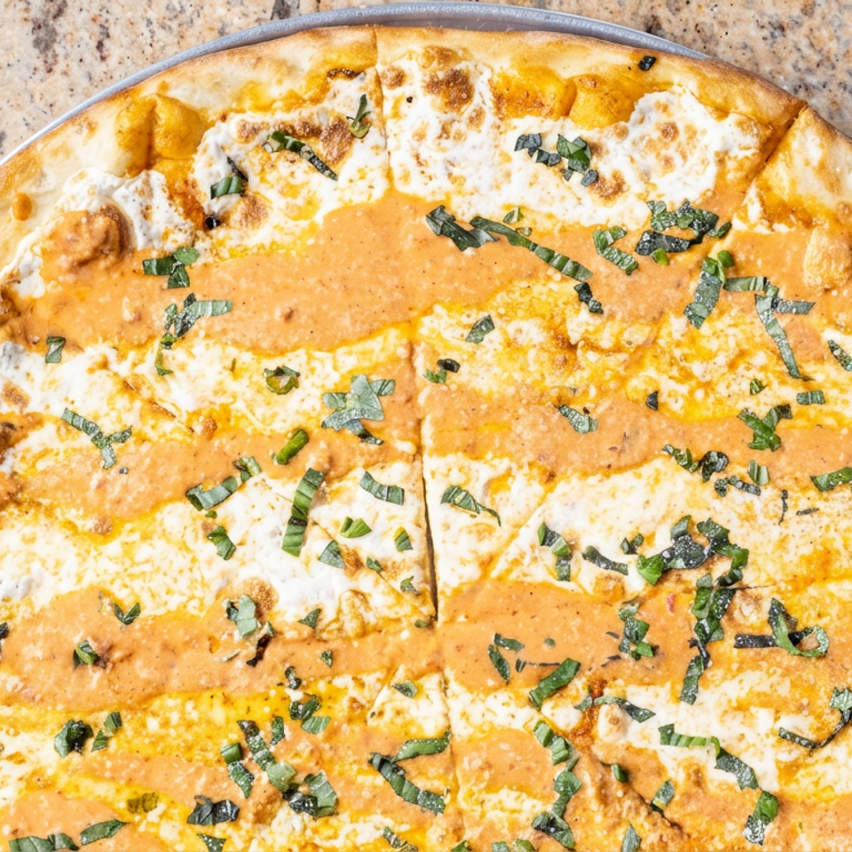 Overhead view of a thin-crust pizza topped with melted cheese, creamy orange sauce, and fresh basil on a speckled countertop.