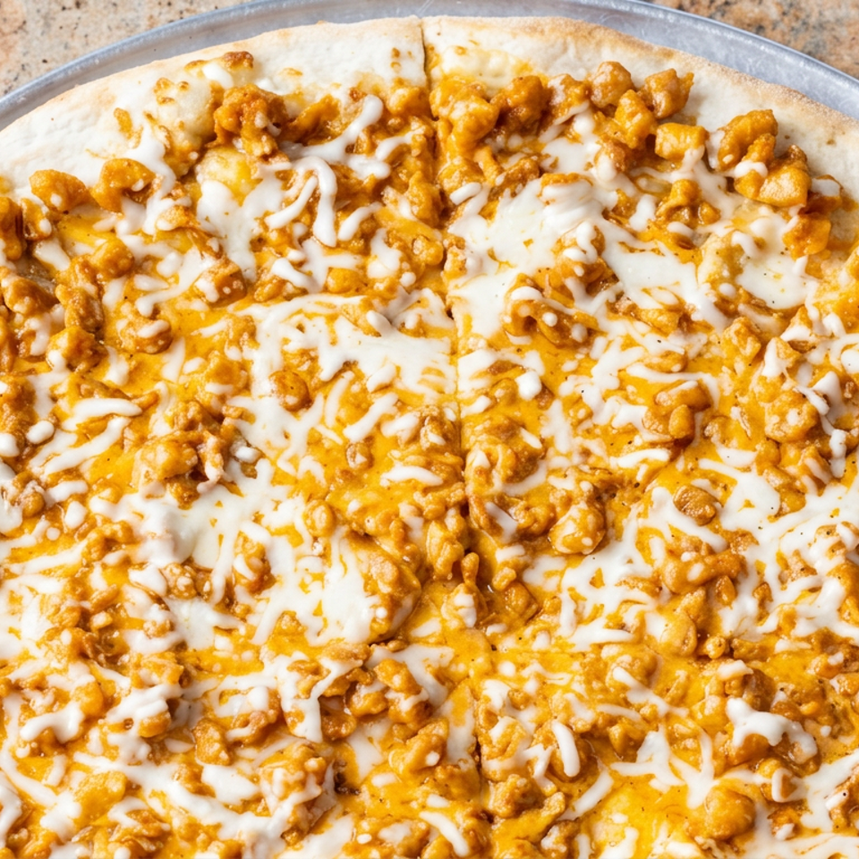 Close-up of a cheesy pizza topped with seasoned chicken pieces and drizzled sauce on a metal serving tray.